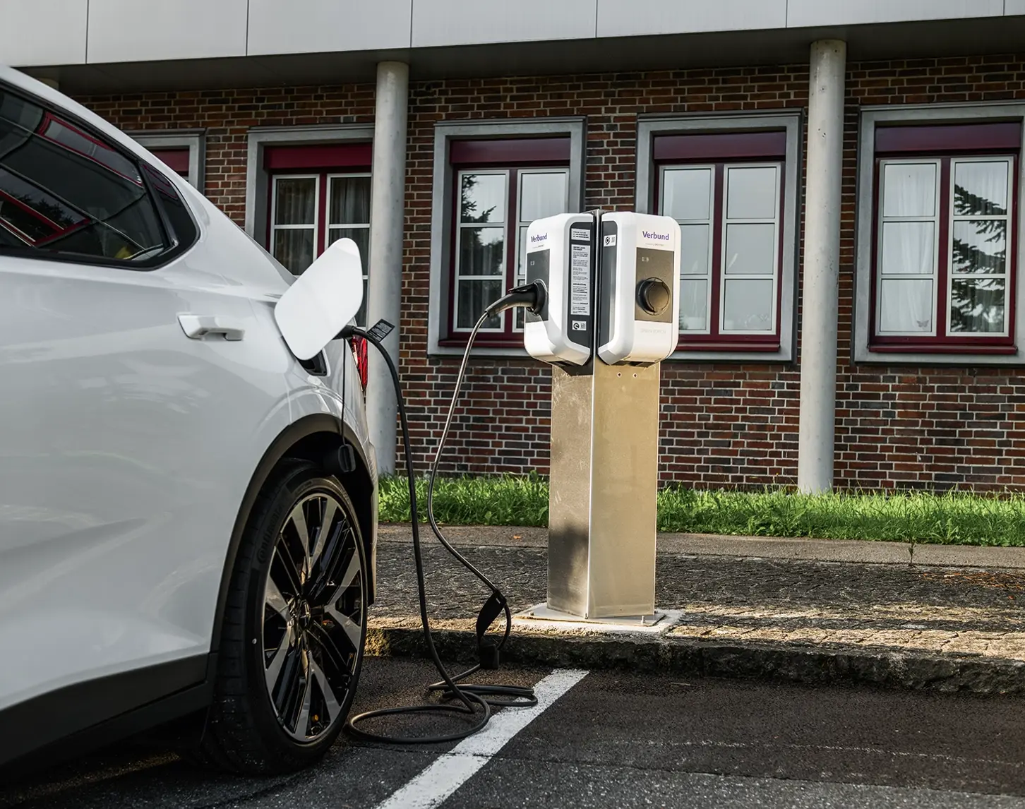 Man sieht eine Nahaufnahme von einem weißen Auto vor eine E-Ladestation. Es wird gerade geladen. Im Hintergrund sieht man ein Industriegebäude.