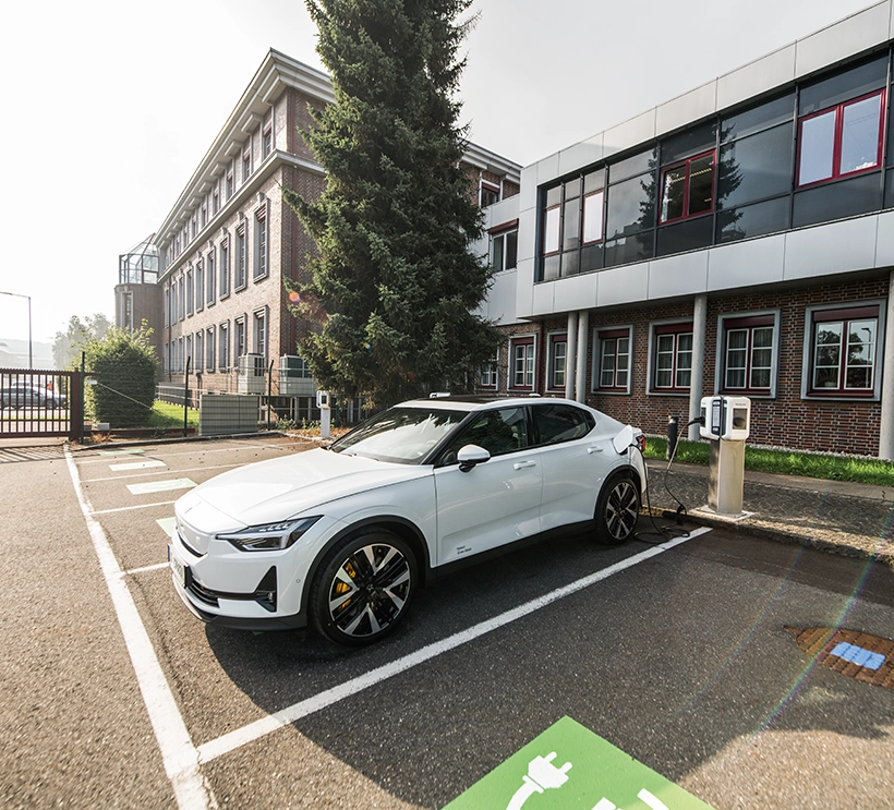 Ein weißes Auto parkt vor eine E-Ladestation. Es wird gerade geladen. Im Hintergrund sieht man ein Industriegebäude.