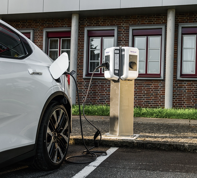 Man sieht eine Nahaufnahme von einem weißen Auto vor eine E-Ladestation. Es wird gerade geladen. Im Hintergrund sieht man ein Industriegebäude.