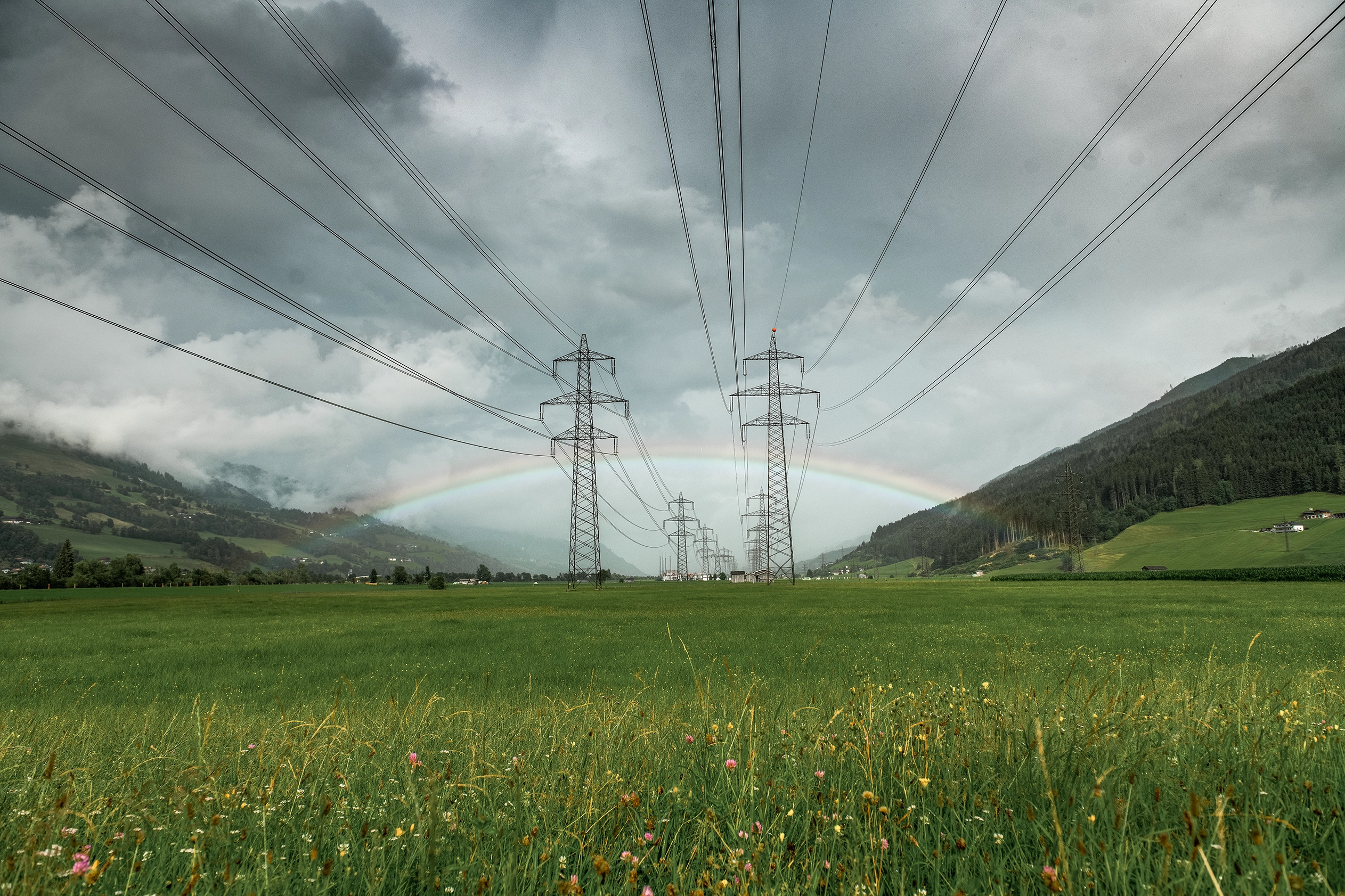 Im Vordergrund zeigt sich eine grüne Blumenwiese. Im Mittelgrund sieht man Strommasten und im Hintergrund zeigen sich Wolken und ein Regenbogen.