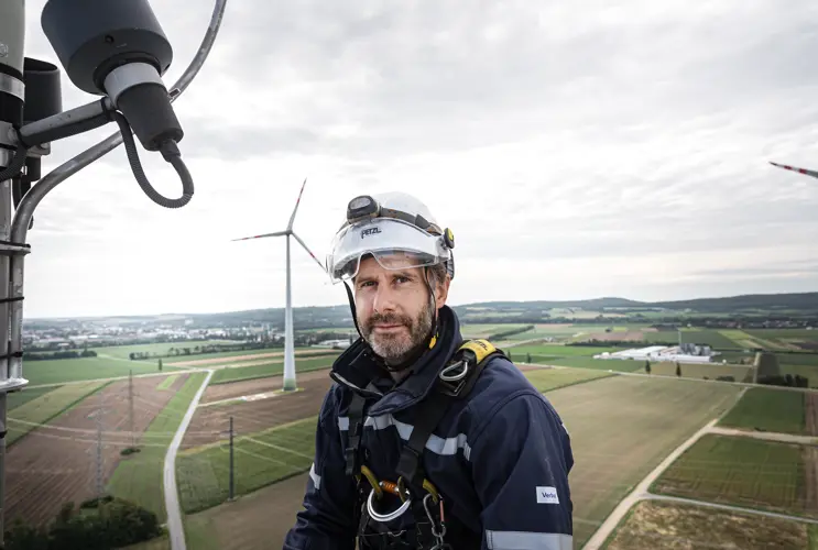 Ein VERBUND-Mitarbeiter in Sicherheitskleidung, Sicherung und Helm blickt in die Kamera. Er befindet sich auf einem Windrad.