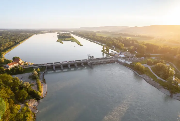 Ein Blick von oben auf das Wasserkraftwerk Ering-Frauenstein im Süden von Deutschland. Das Kraftwerk ist umringt von Wiesen und Wäldern. Die Sonne geht am rechten Bildrand unter.