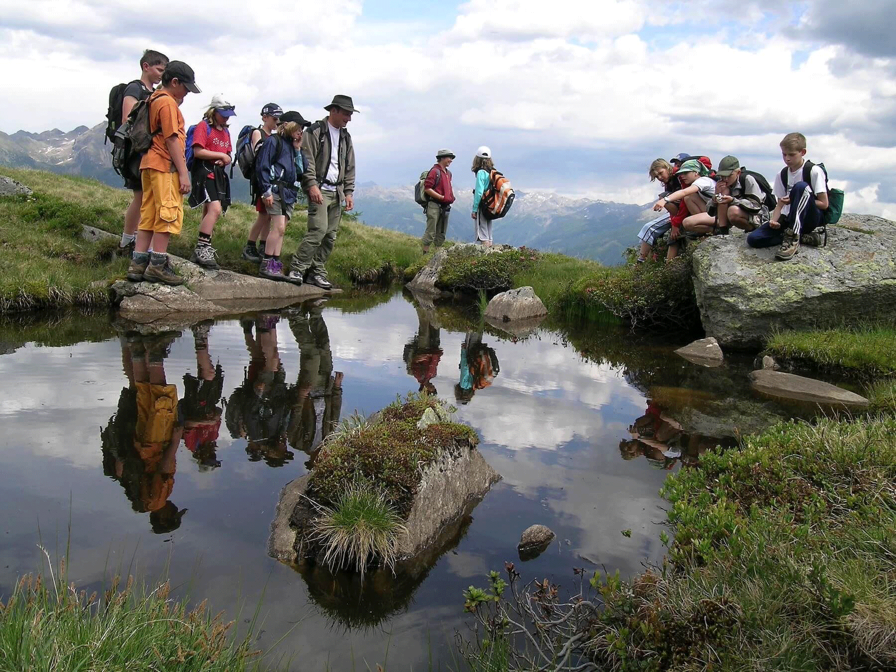 Wir sehen eine Gruppe Kindern mit einem Erwachsenen in den Bergen. Die Wiesen sind grün und der Himmel ist strahlend blau.