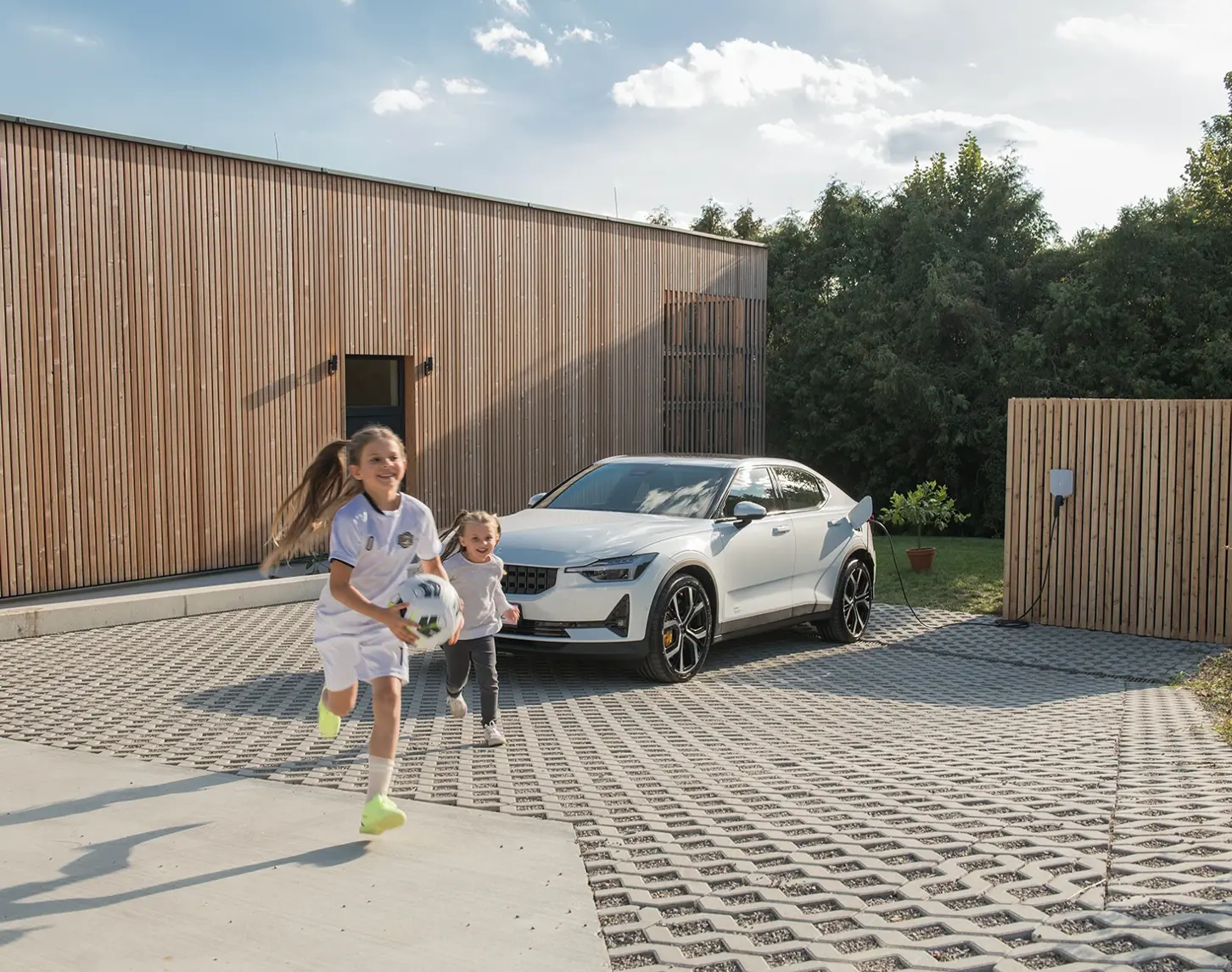 Ein Schwesternpaar läuft freudig mit einem Fussball von einem modern gestalteten Holzhaus auf den Fotografen zu. Im Hintergrund sieht man ein weißes E-Auto, dass gerade aufgeladen wird.