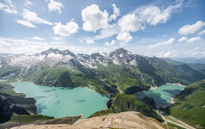 Der Kapruner Stausee mit seinen Gebirgen im Hintergrund bei strahlenden Sonnenschein aus der Vogelperspektive.