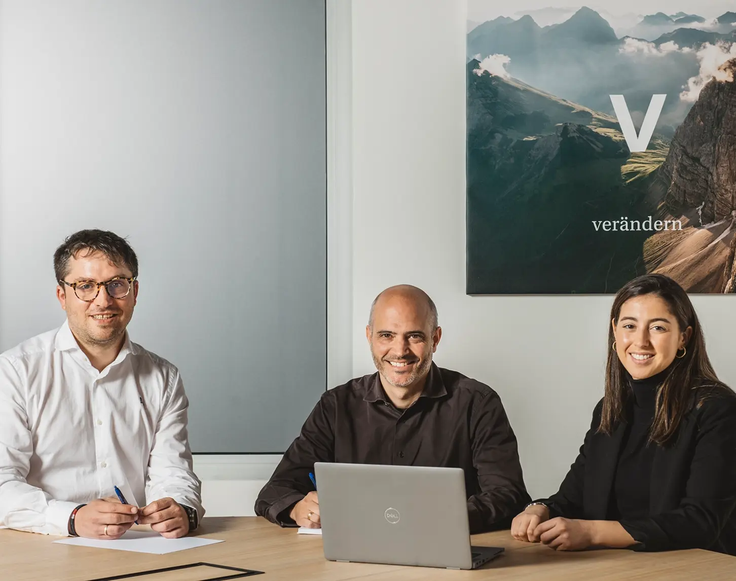 Drei VERBUND-Mitarbeiter:innen sitzen in einem Konferenzraum vor einem Lap-Top und lächeln in die Kamera.