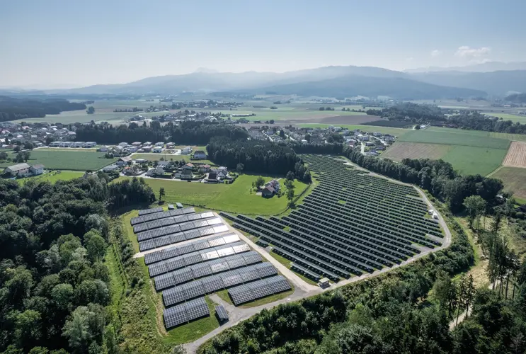 Ein Blick von oben auf eine großflächige Photovotaikanlage. Der Solarpark ist von sattem grün umgeben. Im Hintergrund sieht man kleinere malerische Ortschaften.