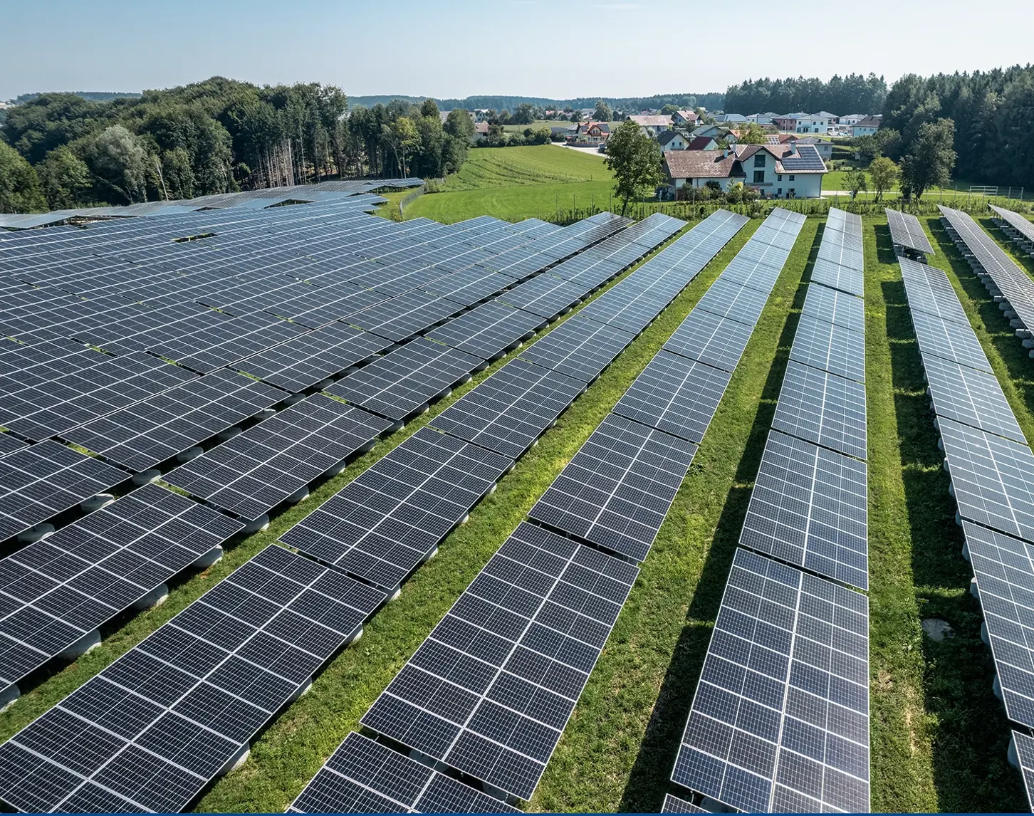 Ein Blick von oben auf eine großflächige Photovotaikanlage. Der Solarpark ist von sattem grün umgeben. Im Hintergrund sieht man kleinere malerische Ortschaften.