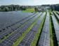 Ein Blick von oben auf eine großflächige Photovotaikanlage. Der Solarpark ist von sattem grün umgeben. Im Hintergrund sieht man kleinere malerische Ortschaften.