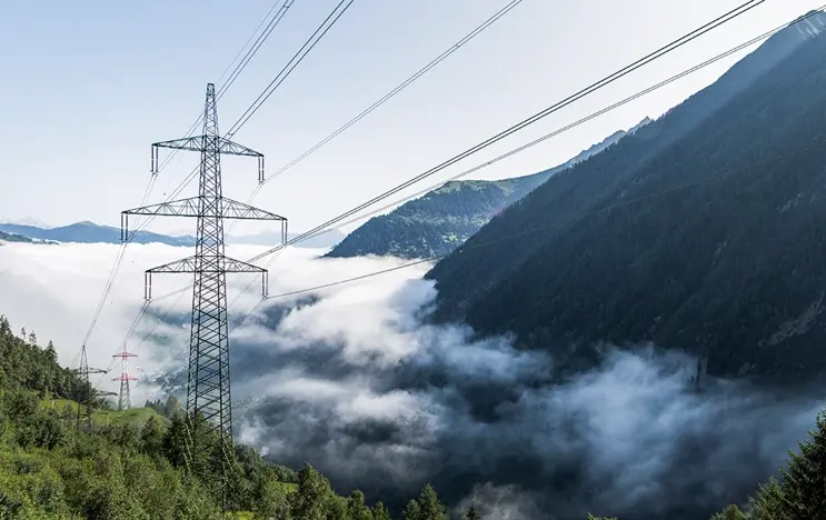 Wir blicken von oben hinab in eine wolkenverhangenen Tal. Im Mittelpunkt sieht man einen Strommast. Die Wälder recht uns links davon sind grün.