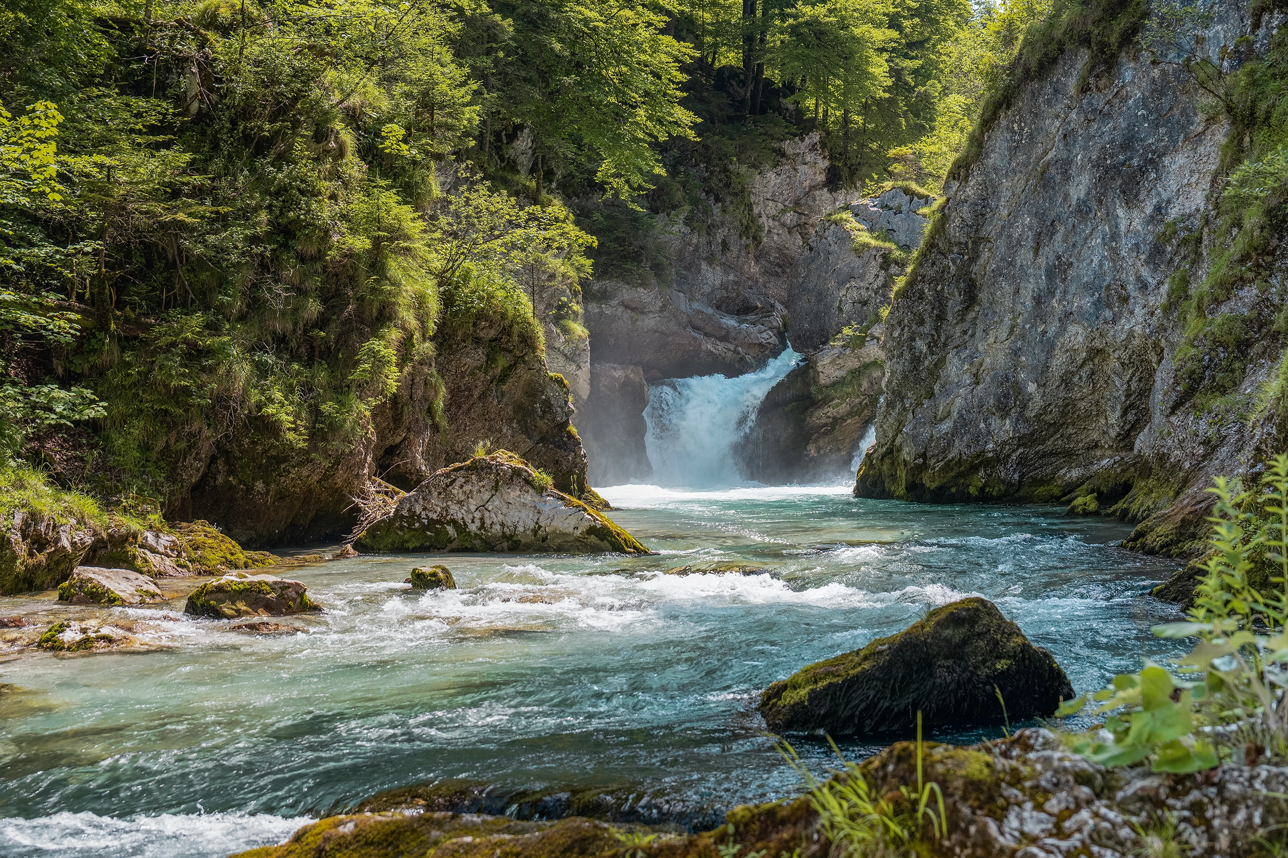 Ein strahlender blauer Bergbach schlängelt sich durch eine Klamm. Die Sonne scheint und zartes Grün ist recht und links sichtbar.