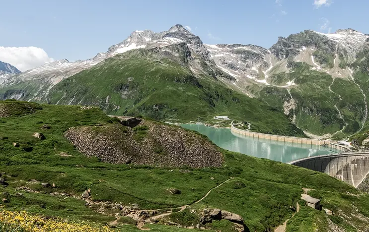 Im Mittelpunkt befindet sich der Stausee bei Kaprun. Im Vordergrund sehen wir Wiesen mit vielen gelben Blumen im Hintergrund türmen sich die Bergwelten auf.