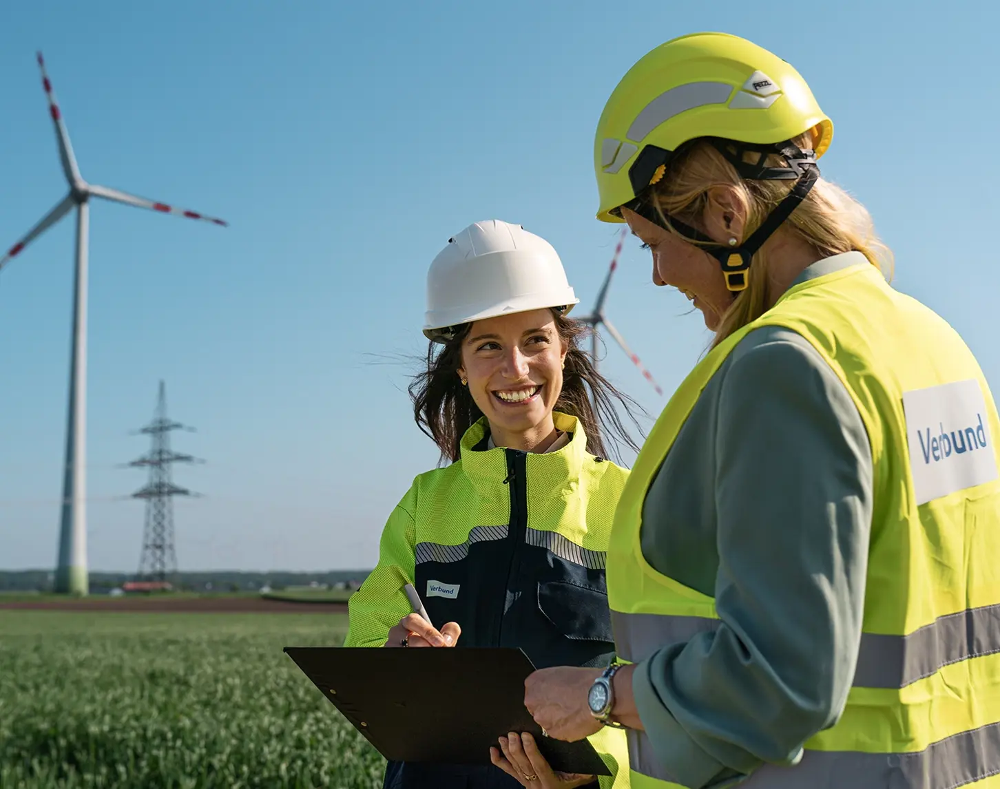 Zwei VERBUND-Mitarbeiterinnen besprechen sich. Im Hintergrund sieht man einen Windpark bei Sonnenschein. Beide Frauen tragen Schutzkleidung und Helme.