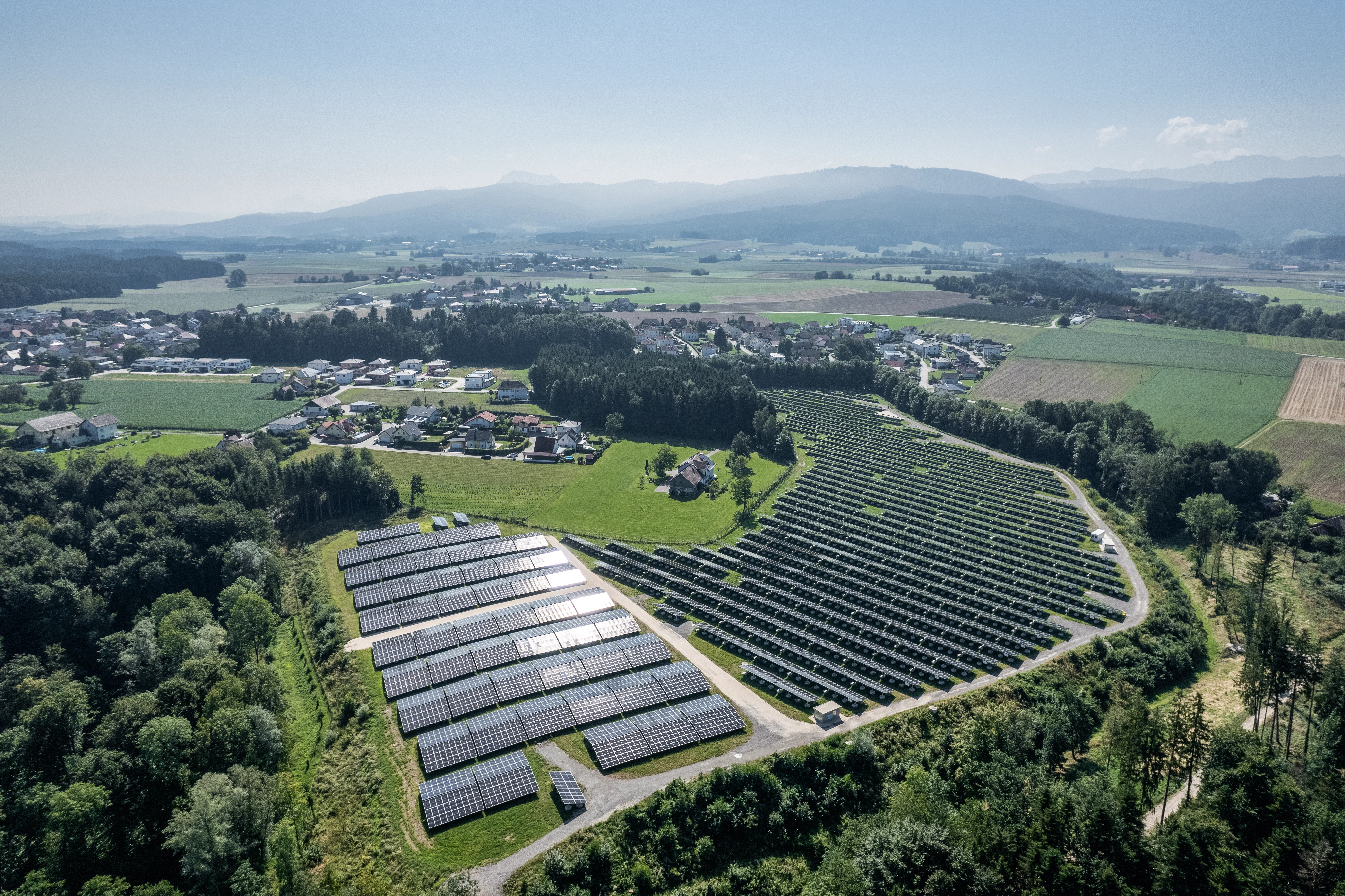 Ein ländliche Photovoltaik-Anlage in ihrem natürlichen Habität. Berger im Hintergrund. Wälder, Wiesen und Dörfer verteilen sich zusätzlich.