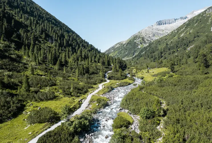 Wir blicken in ein weites Tal mit Wiesen und mächtigen Bergen im Hintergrund. Ein Bergbach schlängelt sich auf uns zu.