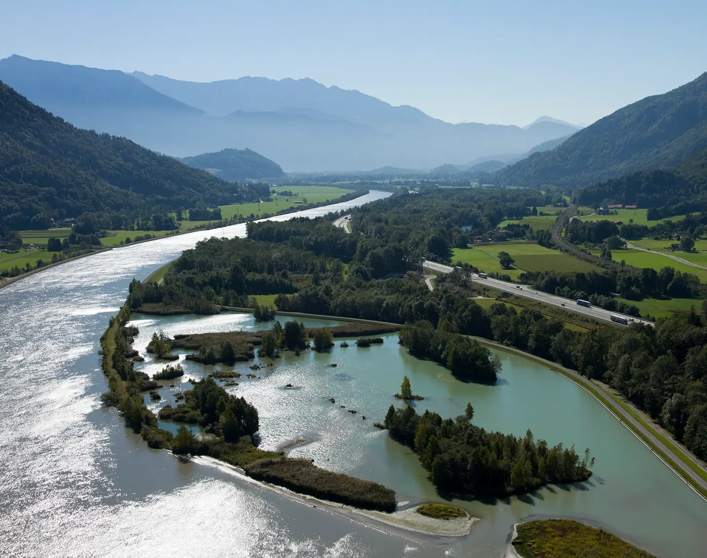 Ein Blick von oben auf den Inn. Das Wasser reflektiert die Sonne. Der Fluss wird begleitet von Wiesen, Wäldern und Bergketten. Der Himmel ist strahlend blau.