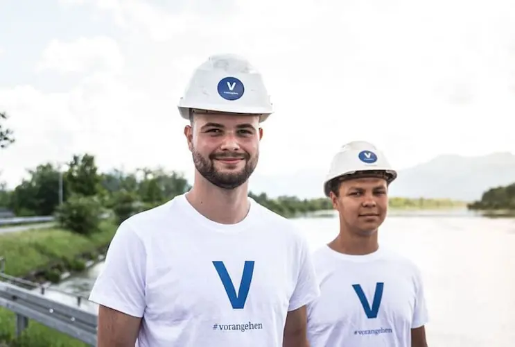 Zwei junge VERBUND-Mitarbeiter in weißen T-Shirts mit blauem V und Helmen lächeln in die Kamera.